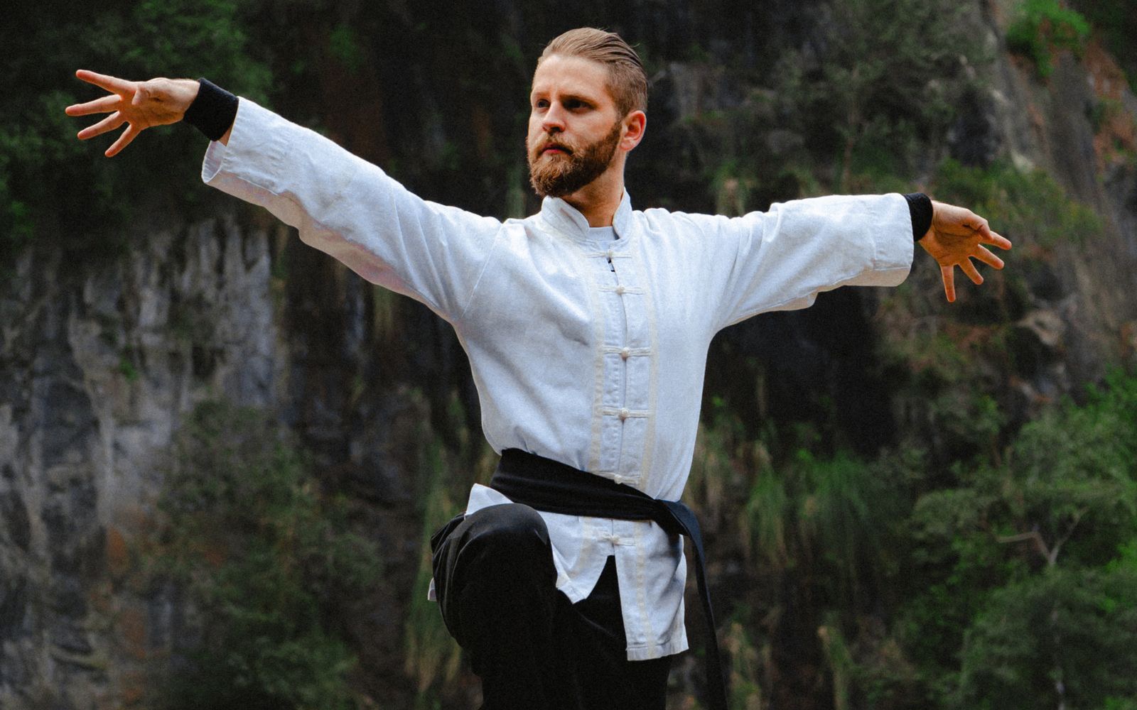 A man in white practising tai chi before a forested cliff